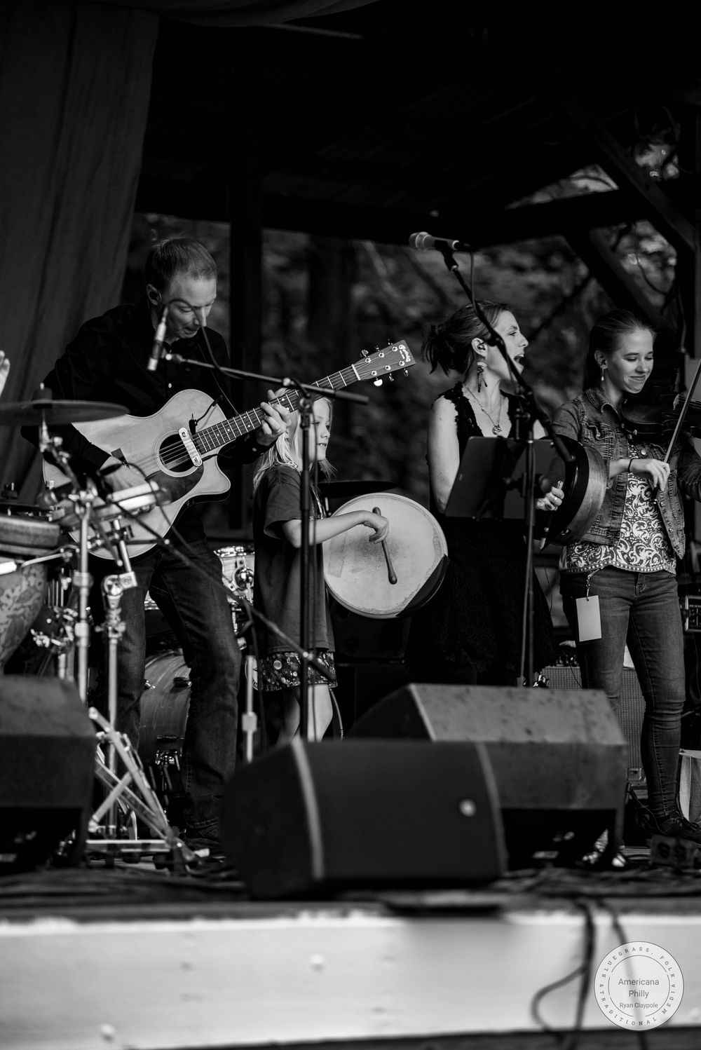 Black and white photo of band performing on outdoor stage with guitarist and frame drum player.