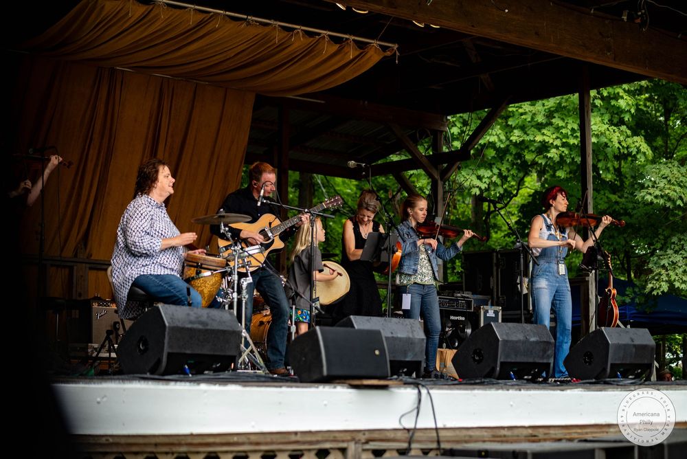 Live band performing on an outdoor stage surrounded by lush green trees at a summer music festival.