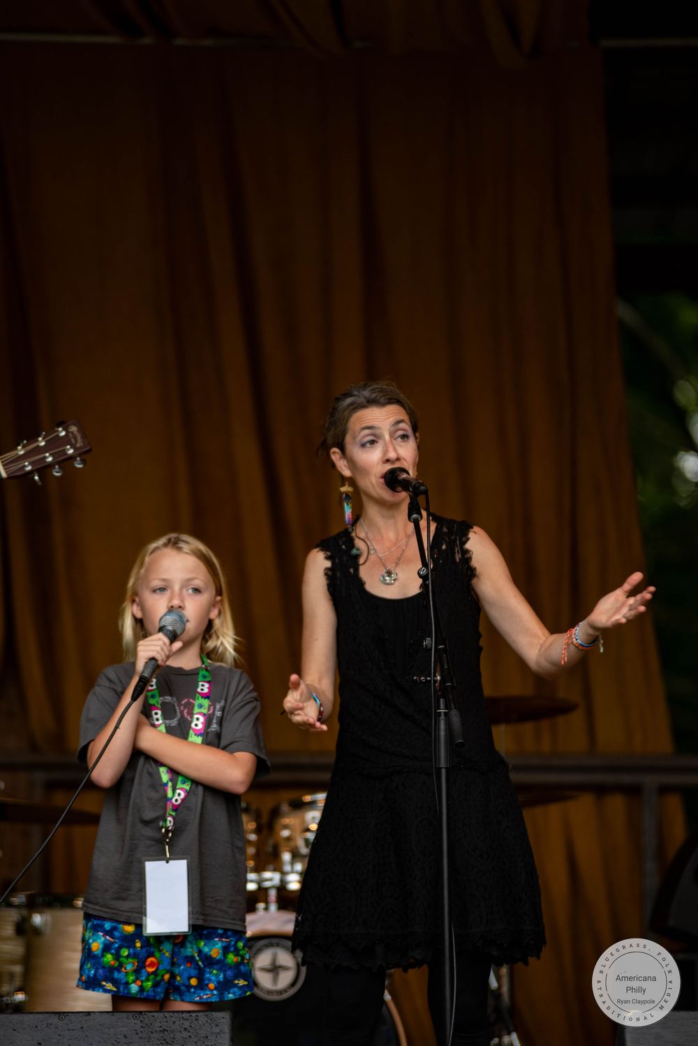 A woman in black dress and young girl sing on stage with microphones at an outdoor performance event.