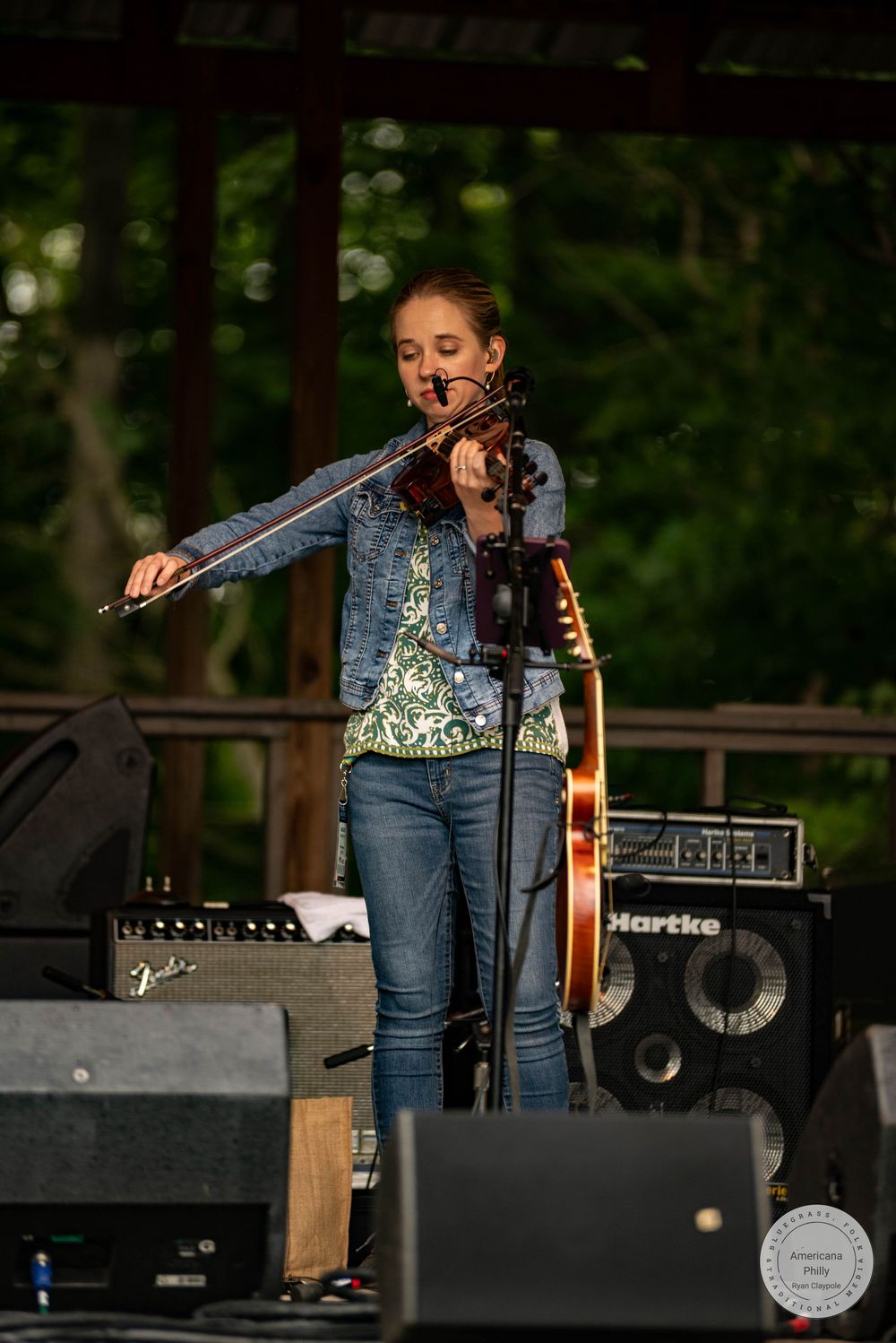 Young woman playing violin on outdoor stage with Fender and Hartke amplifiers, wearing jeans and floral top.