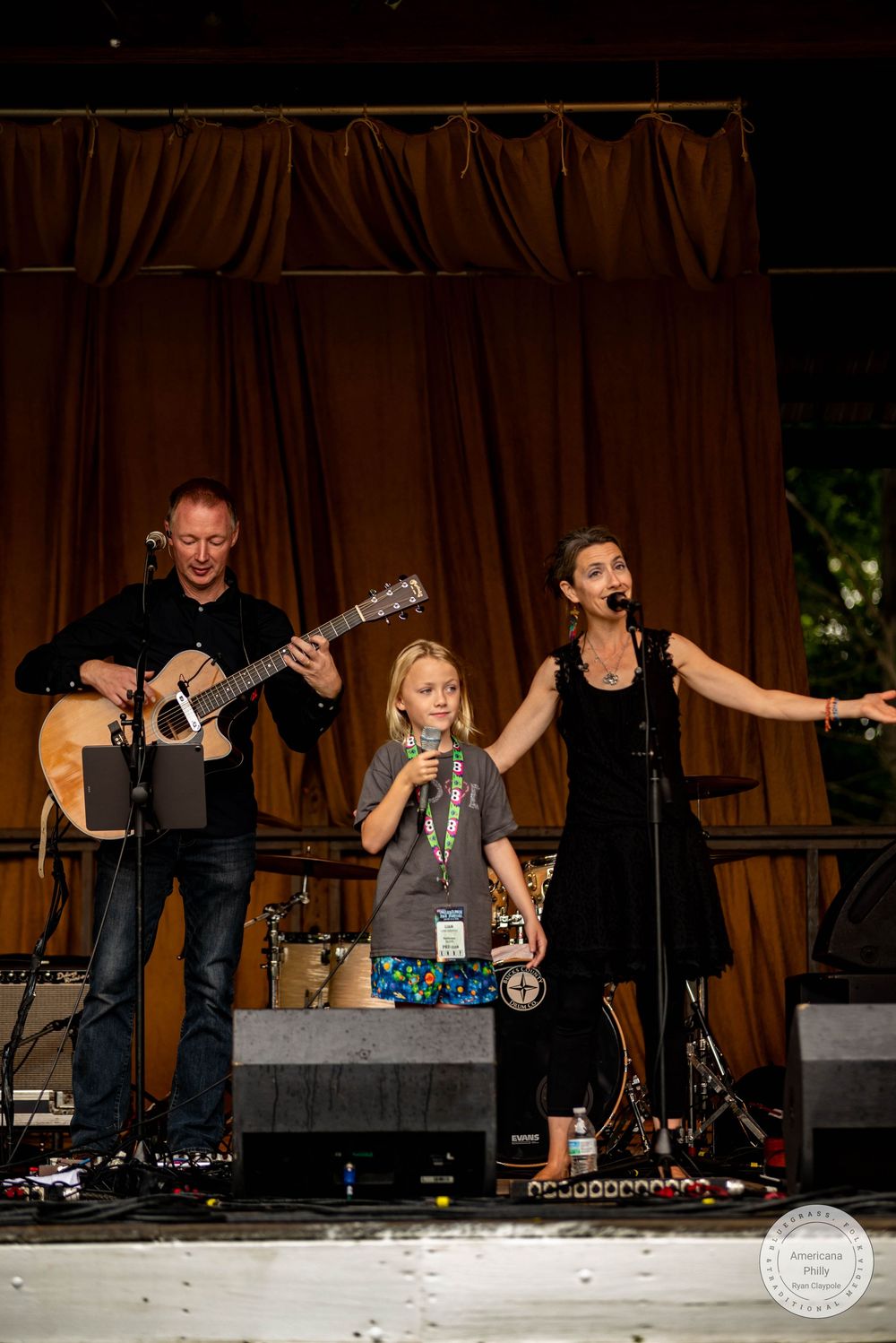 A child sings on stage with a guitarist and female vocalist performing live at an outdoor concert event.