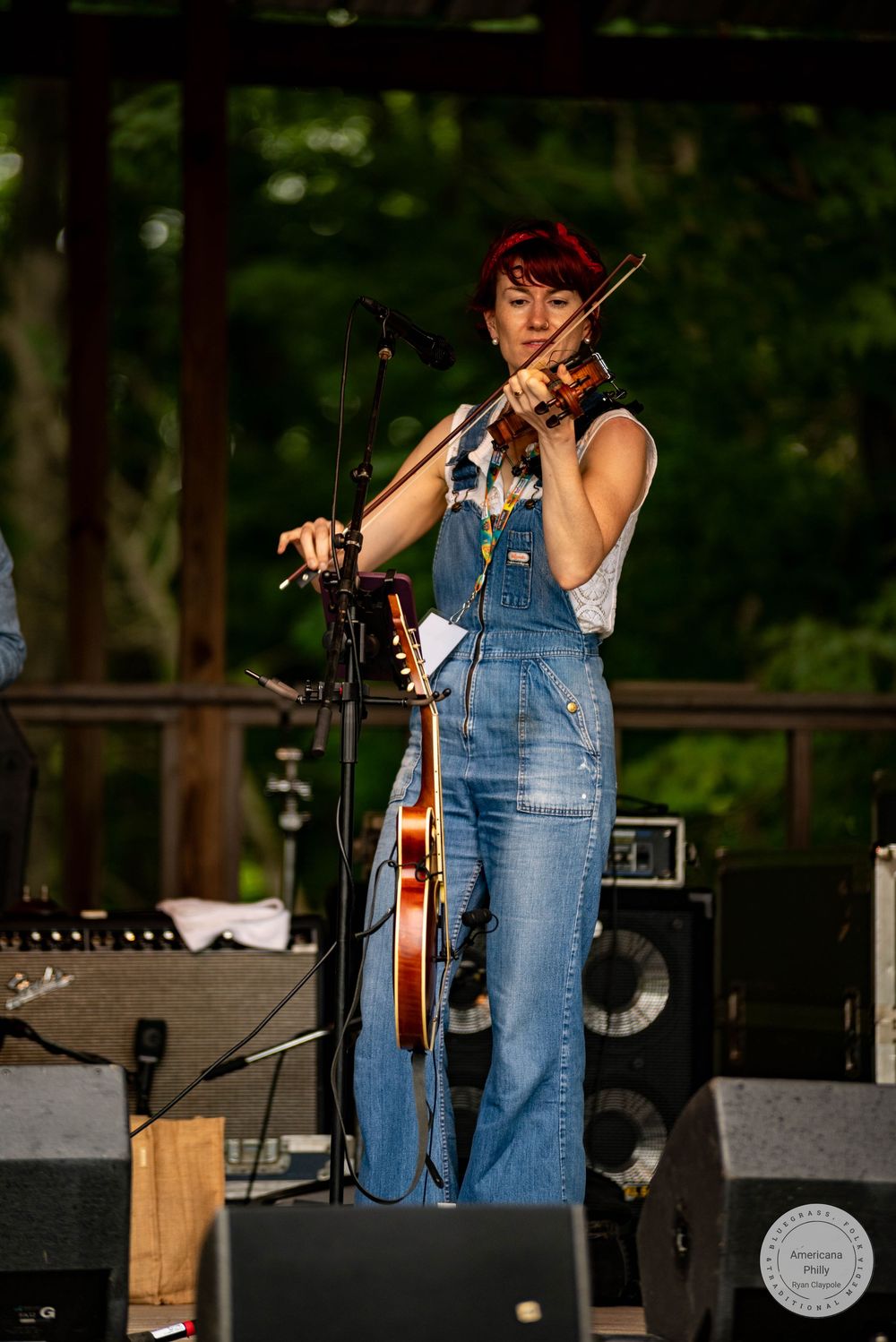 Woman in denim overalls playing violin on outdoor stage surrounded by speakers and microphone stands.