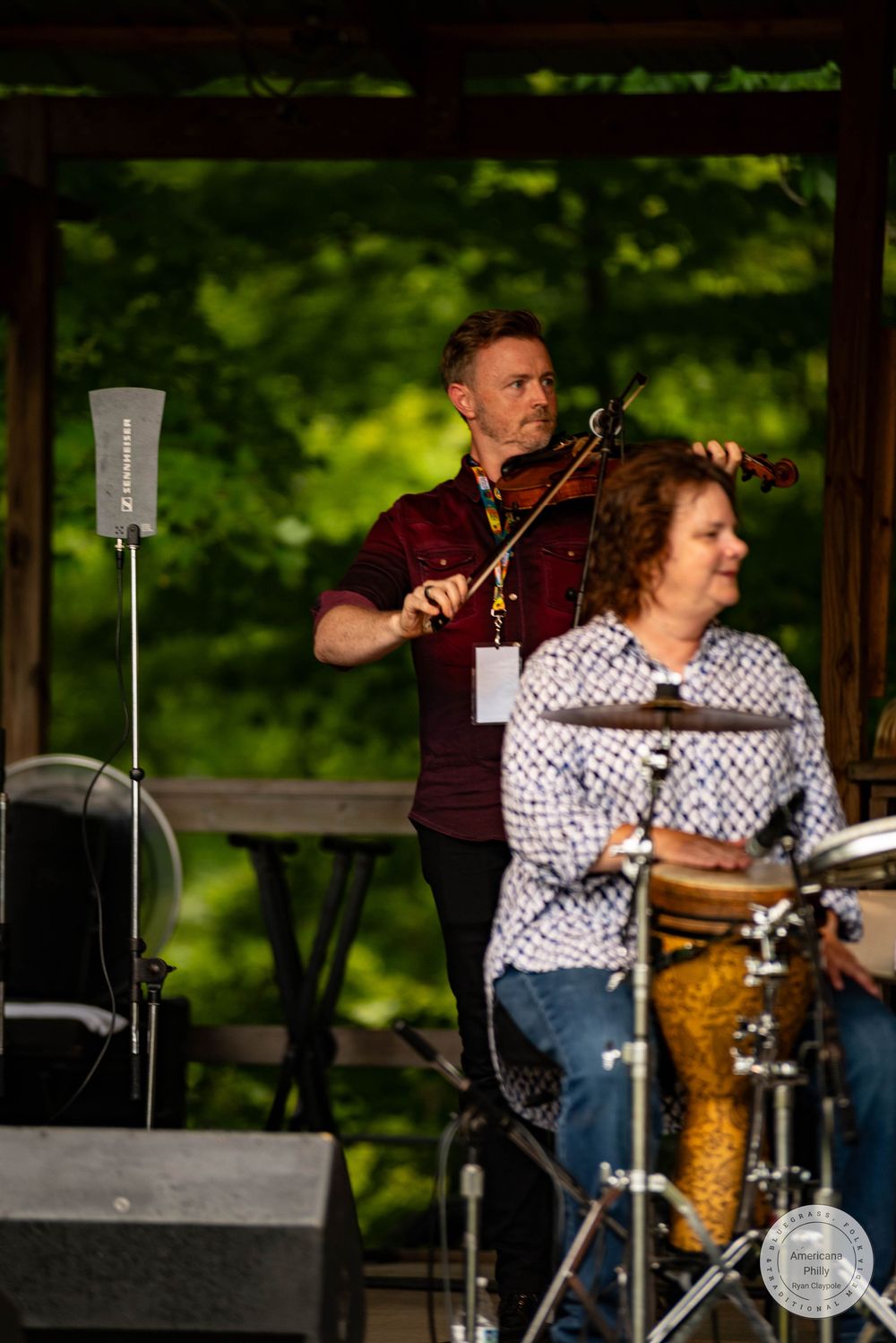 Man playing violin on outdoor stage with woman at drums, performing at a lush green park pavilion.