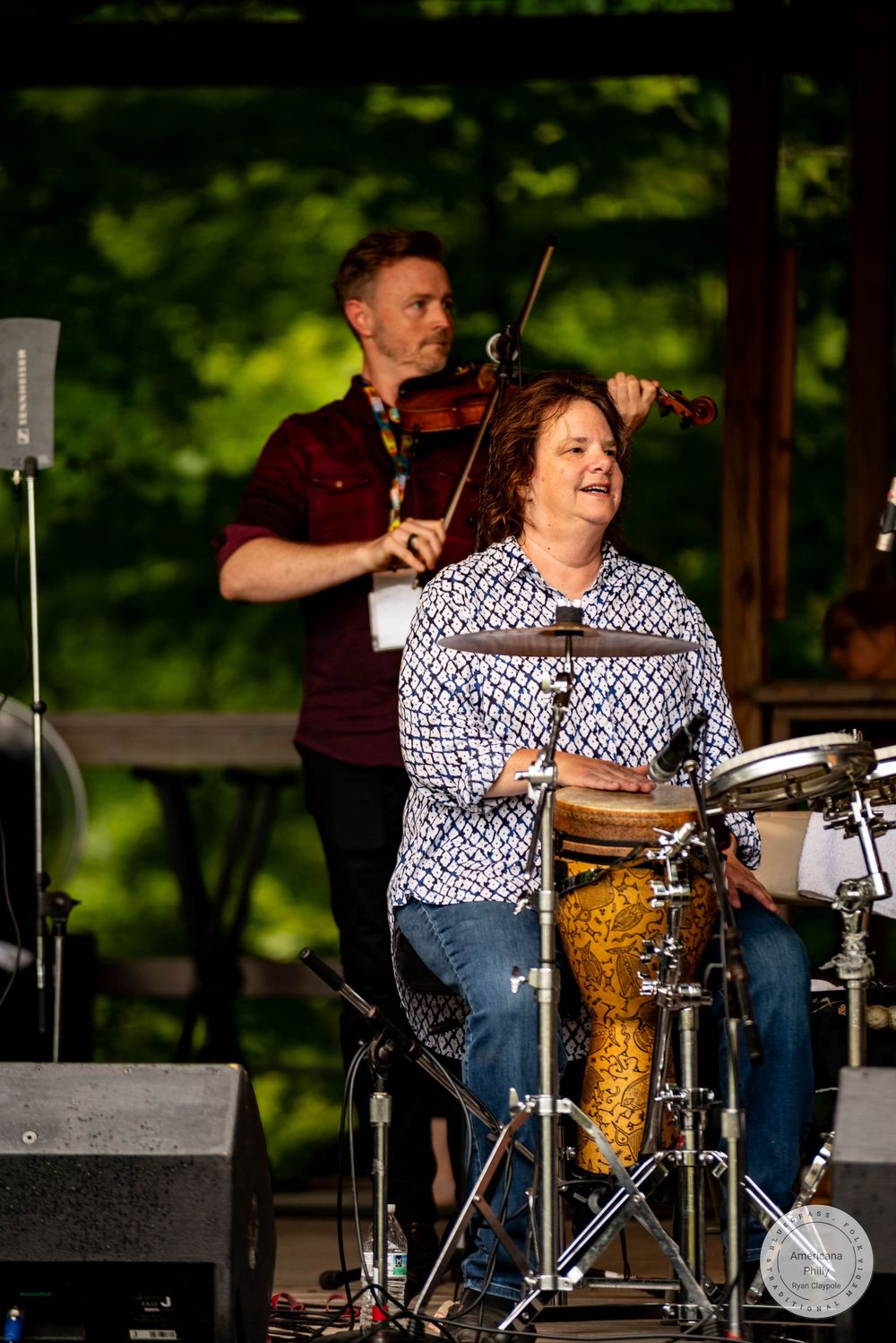 Woman plays drums on outdoor stage while male violinist performs behind her at a live music festival.