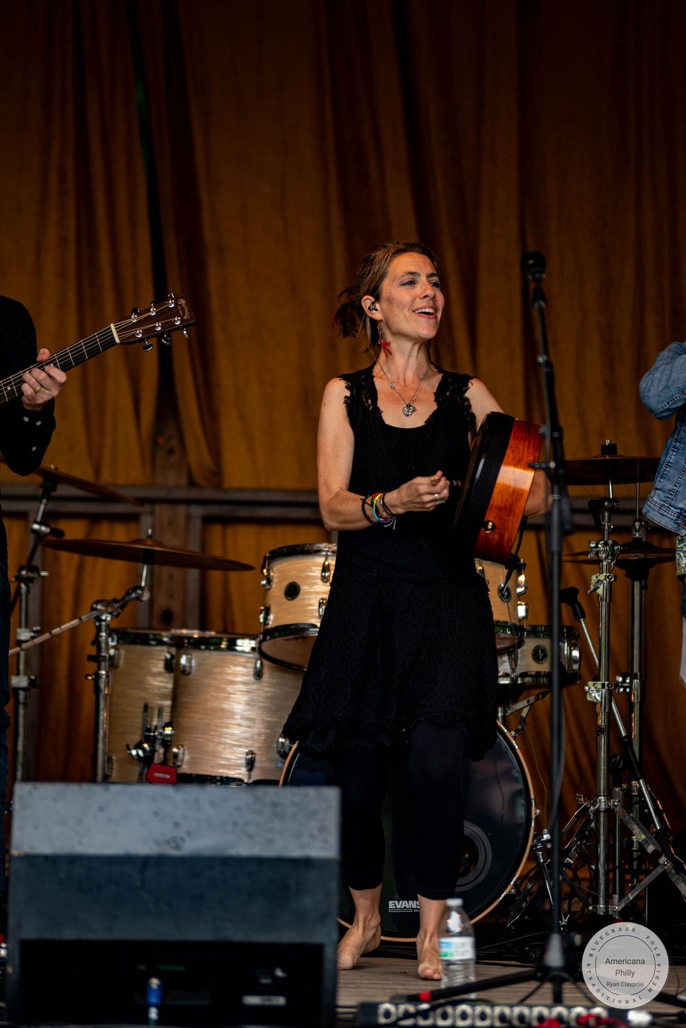 Woman in black dress performs barefoot on stage with guitar and drum kit at an outdoor concert.