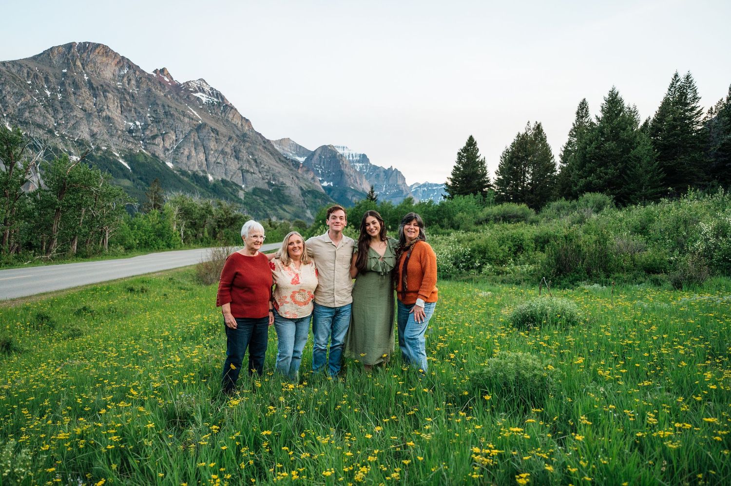 Five people pose together in a lush green meadow with wildflowers, tall pine trees, and dramatic mountain peaks in background.
