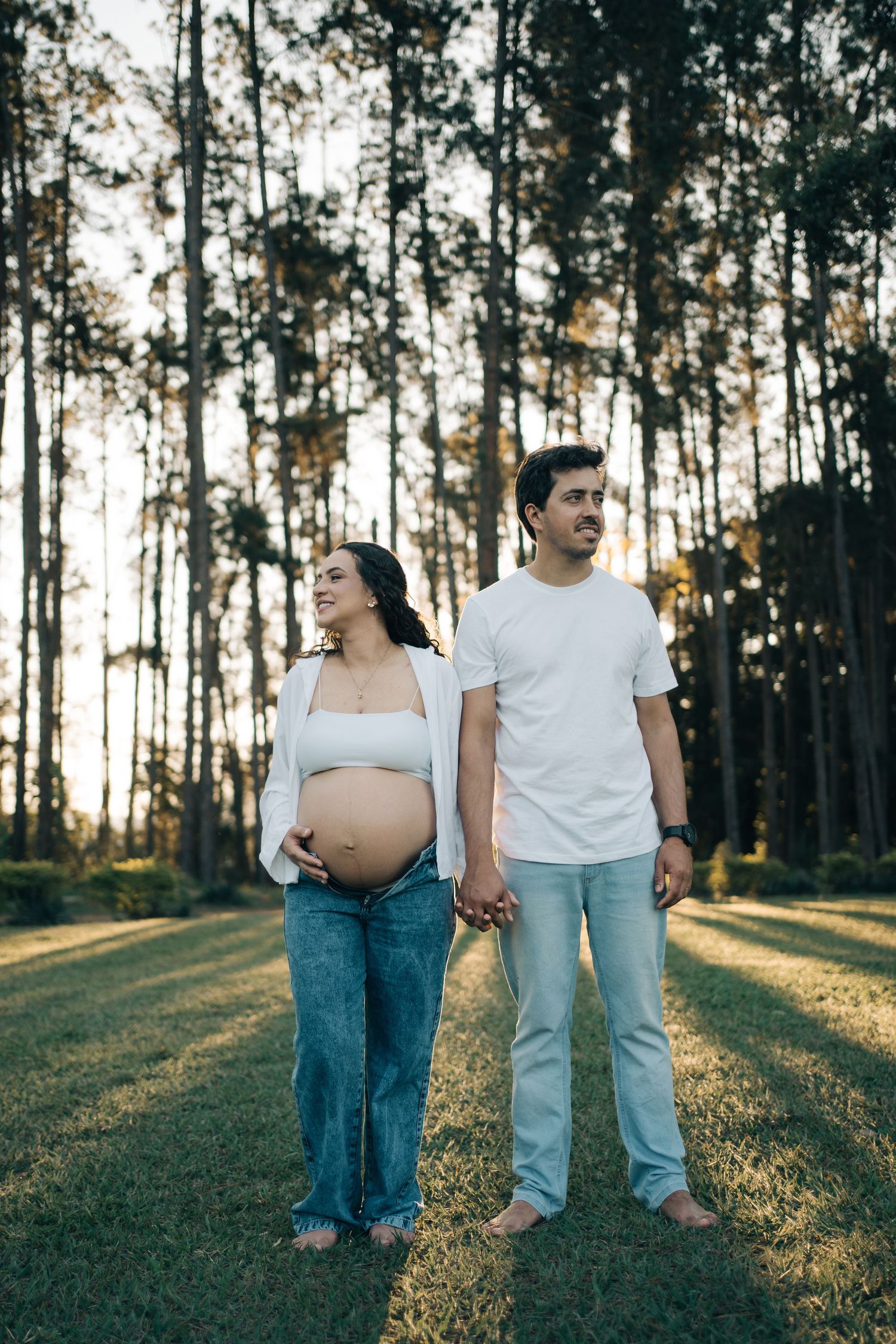 Liliana e Bruno de mãos dadas diante das árvores iluminadas pelo sol na Fazenda Vintém.