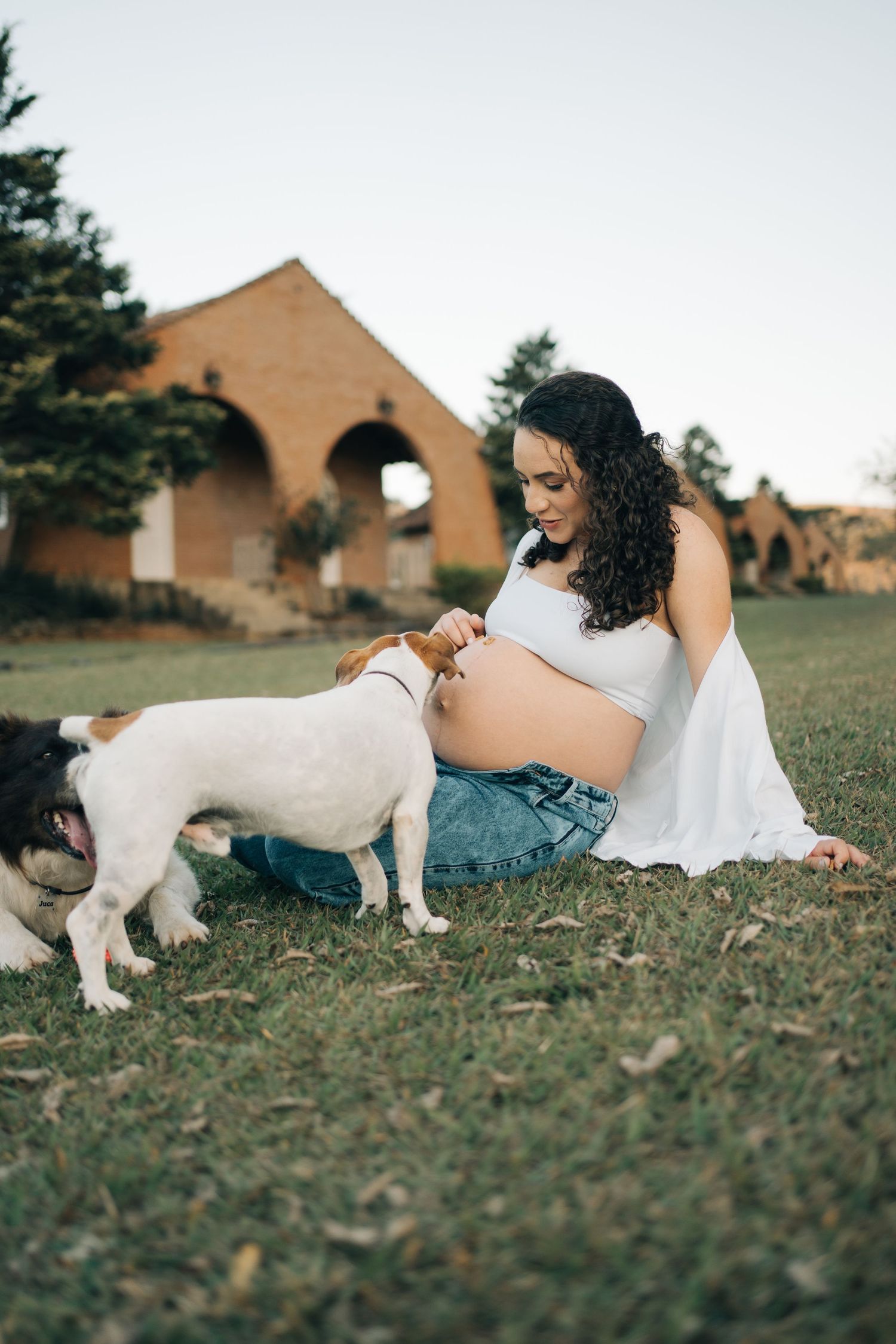 Liliana sentada no gramado com os cachorros próximos à barriga e a Fazenda Vintém ao fundo.