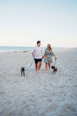 A couple walks their two small dogs along a pristine white sandy beach at sunset.