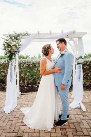 A romantic wedding ceremony under a white pergola decorated with floral arrangements and flowing fabric on a brick patio.