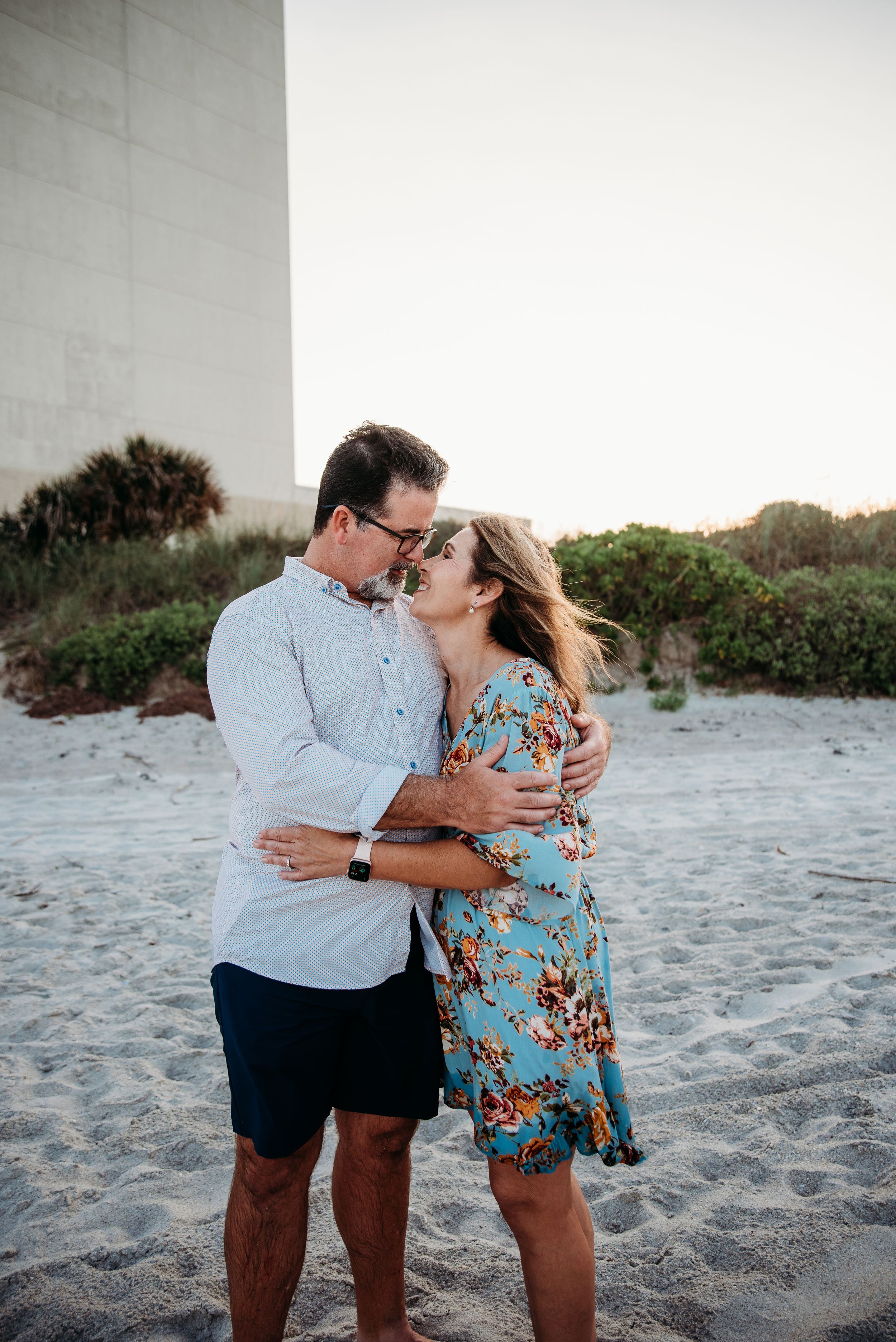 A couple embraces on a sandy beach at sunset, with the woman wearing a blue floral dress and the man in a light shirt.