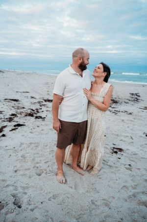 A couple shares a joyful moment together while standing barefoot on a sandy beach at sunset with ocean waves behind them.