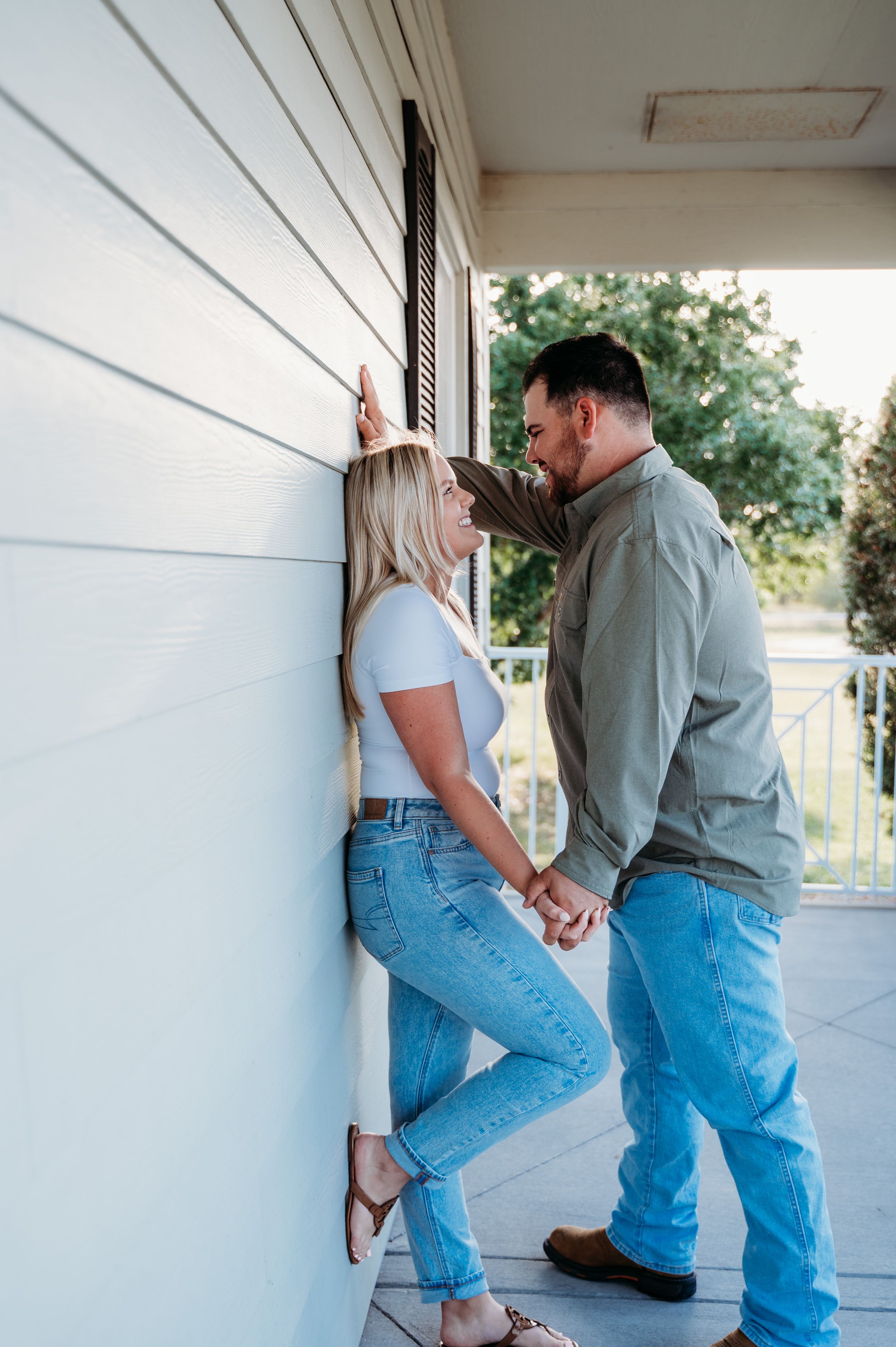 A romantic couple in casual attire share an intimate moment on a covered porch against a white wall.
