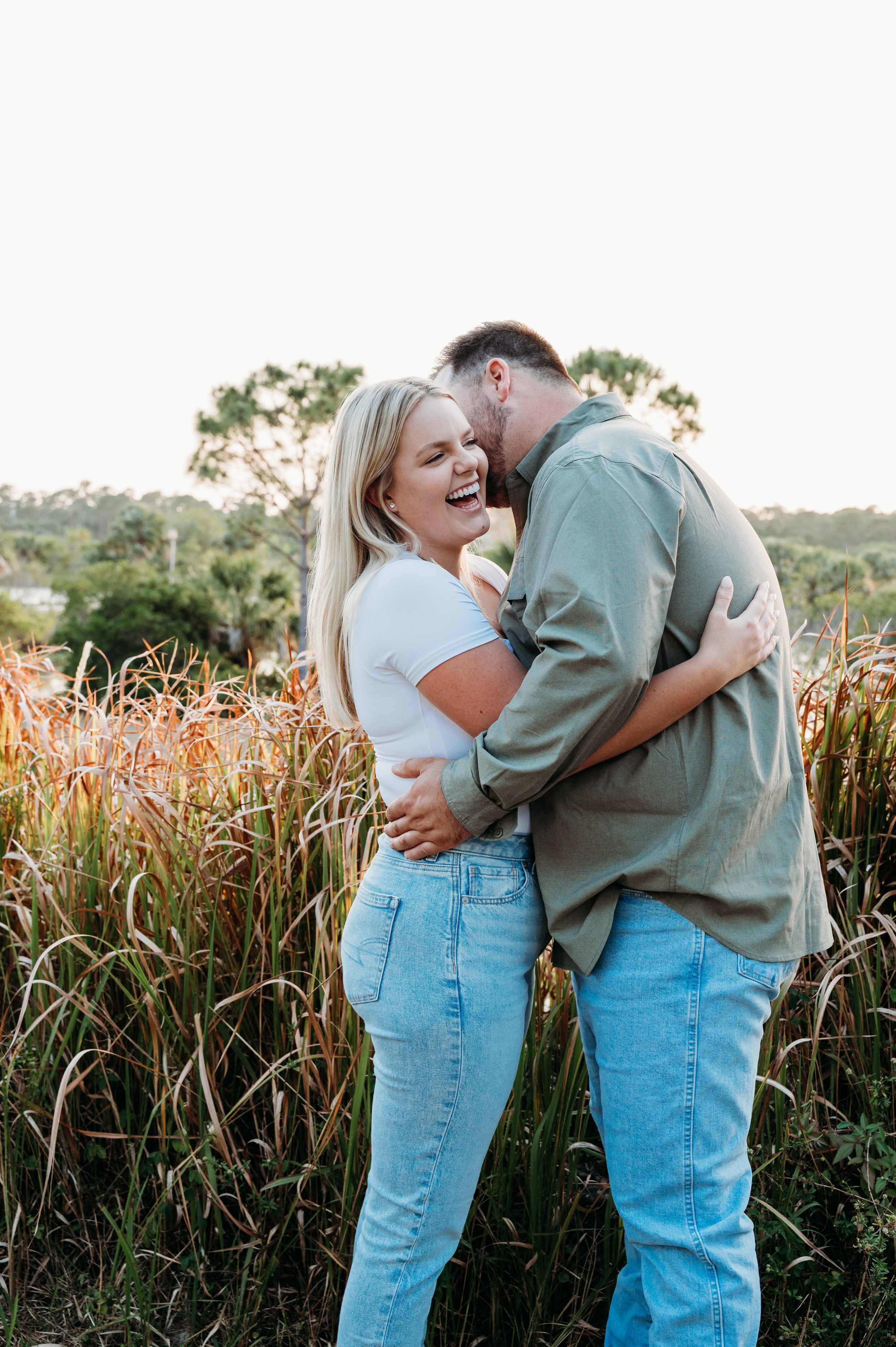 A couple shares a joyful laugh together while embracing in a field of tall grass during golden hour.