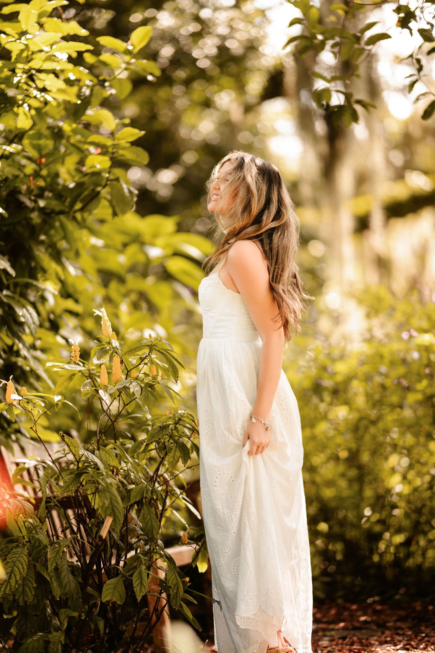 Woman in white maxi dress standing among lush green garden plants in golden sunlight.