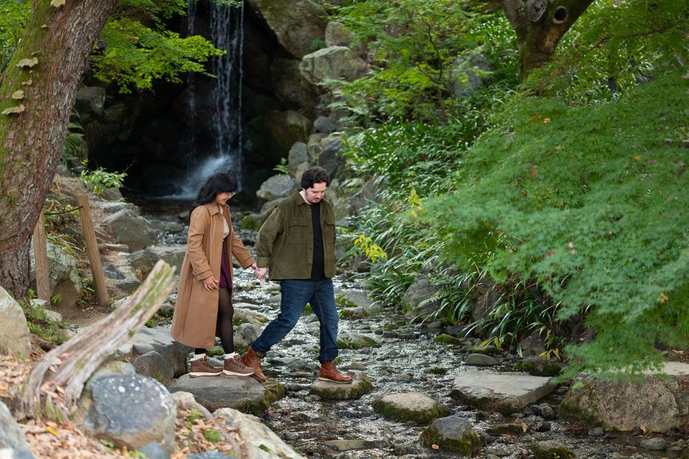Couple holding hands walking on stepping stones beside a serene stream and waterfall in a lush green forest.