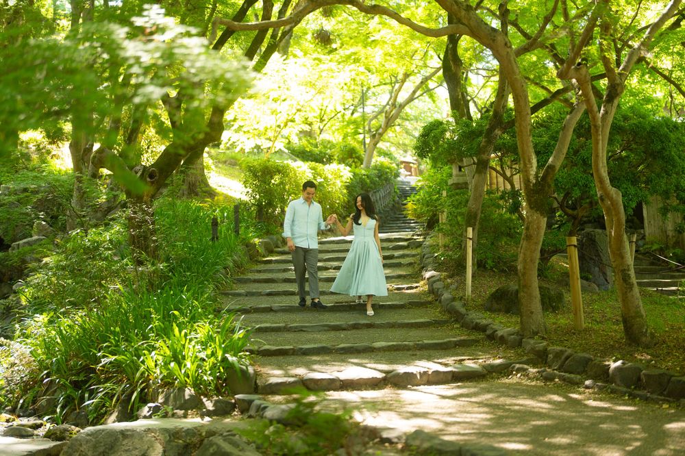 Couple holding hands walking down stone steps in a lush green Japanese garden surrounded by trees.