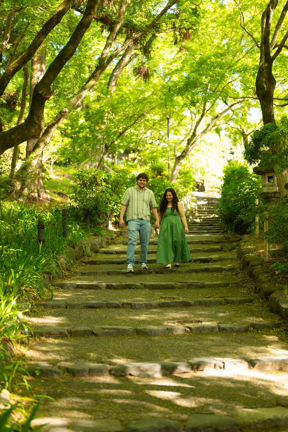 Couple holding hands walking down stone steps in a lush green garden with trees.