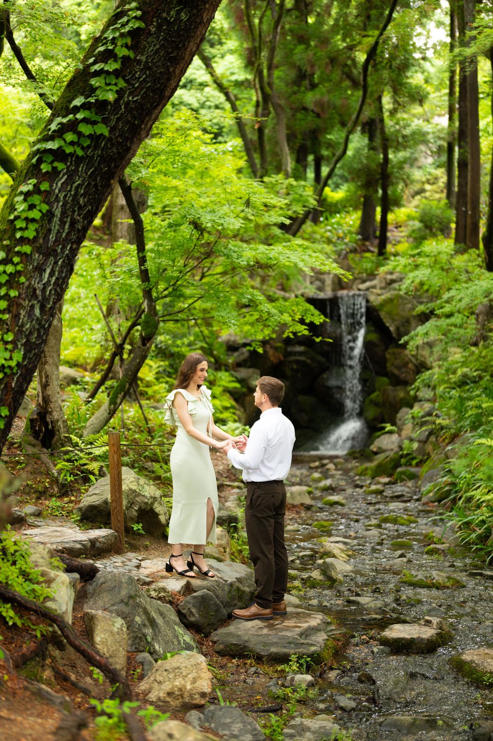 Couple holds hands on stone path by waterfall in lush green forest garden setting.