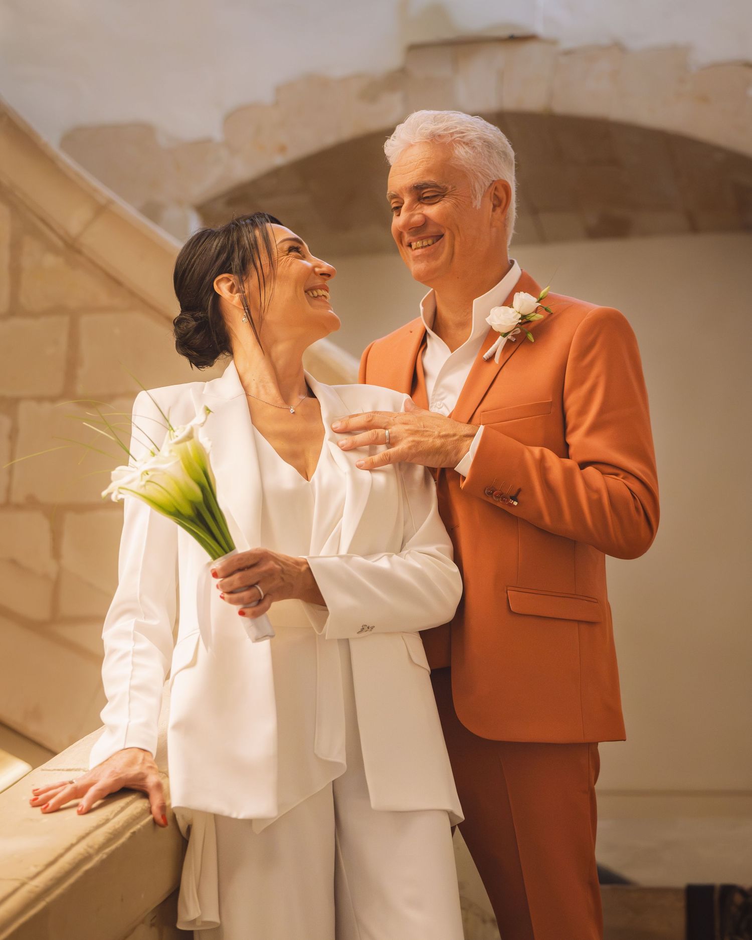 Une mariée en costume blanc tient des callas à côté d'un marié en costume terracotta, tous deux se souriant.