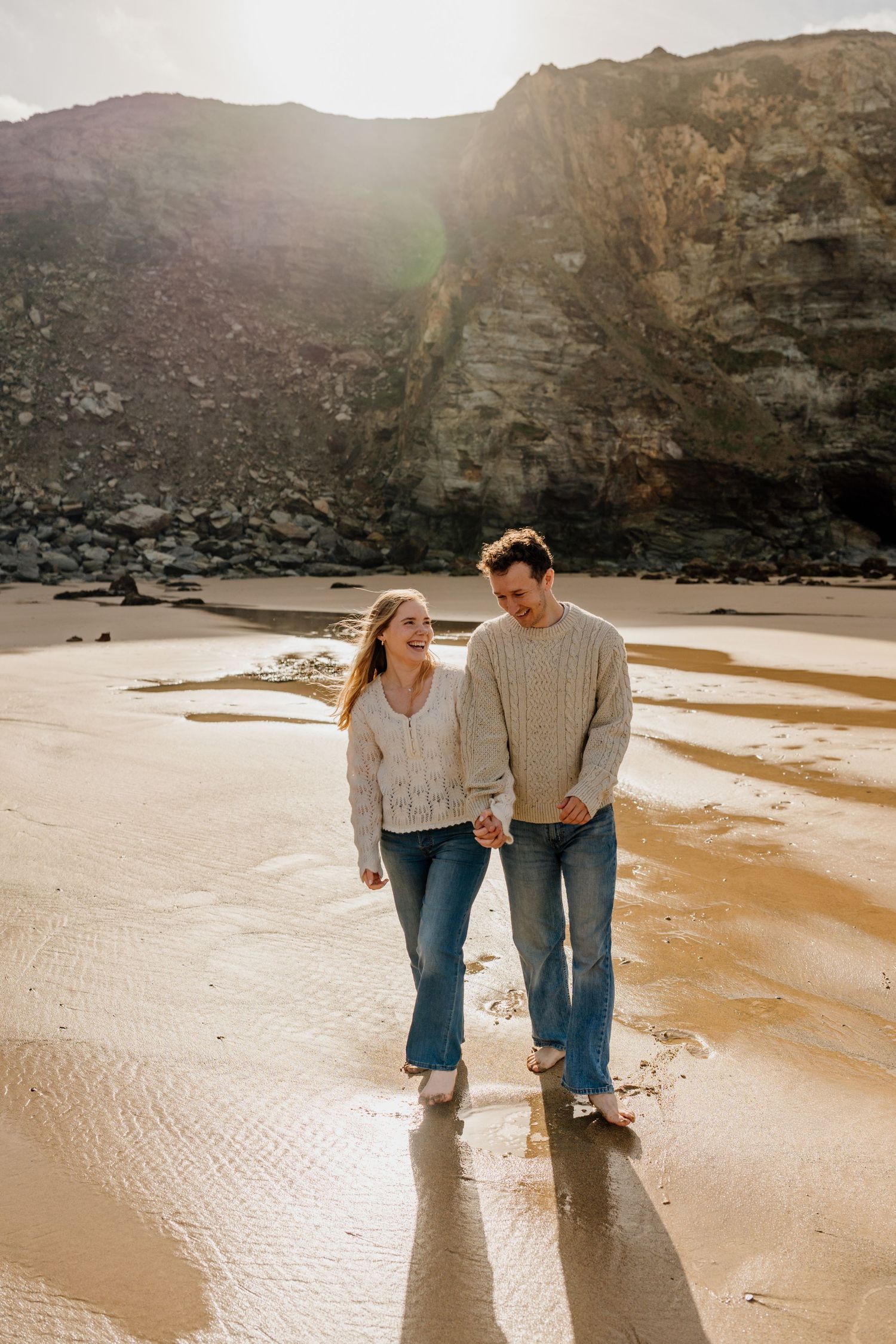 Couple holding hands walking barefoot at watergate bay with dramatic cliffs and sunlight in background.