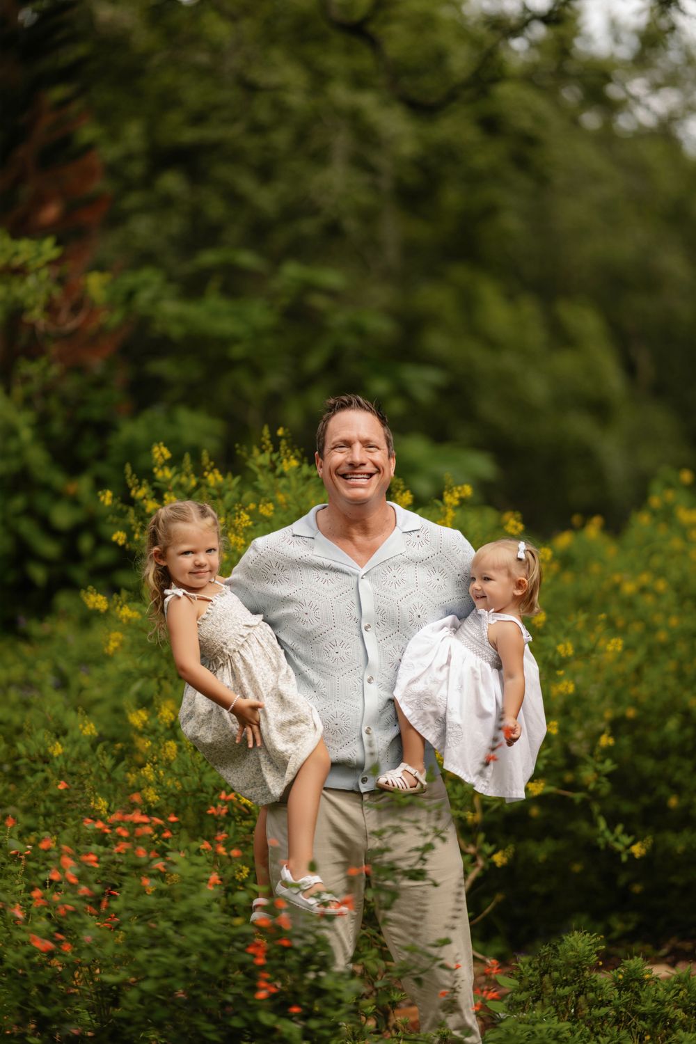 Smiling man in a light blue shirt holds two young girls in white dresses while standing among green foliage and orange wildflowers outdoors.