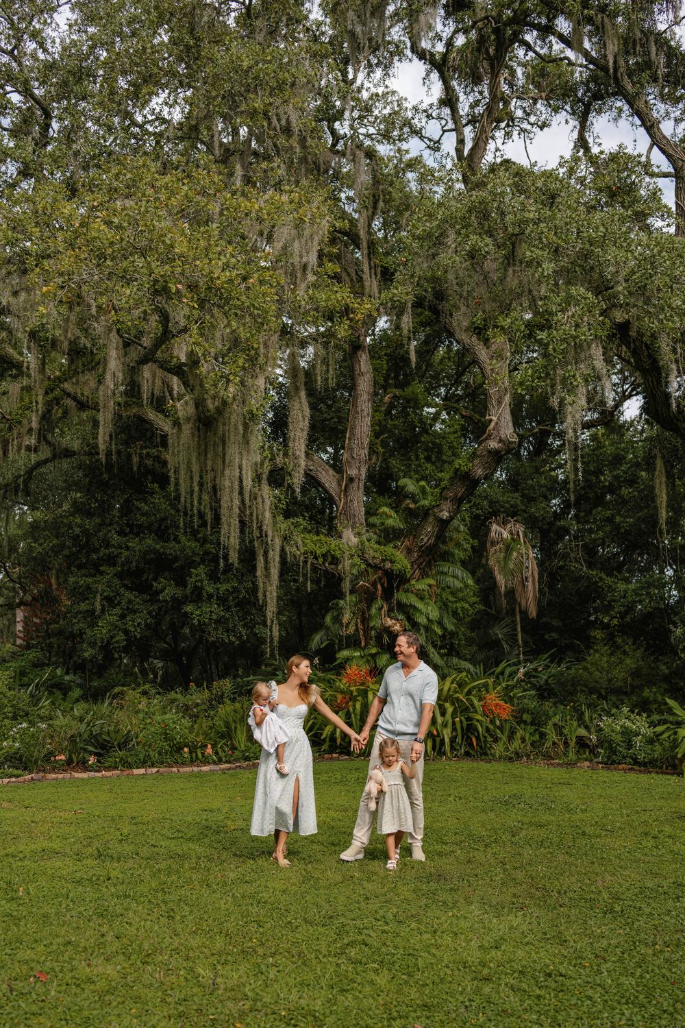 Family of four holding hands on a green lawn beneath large oak trees draped in Spanish moss during an outdoor family portrait session.