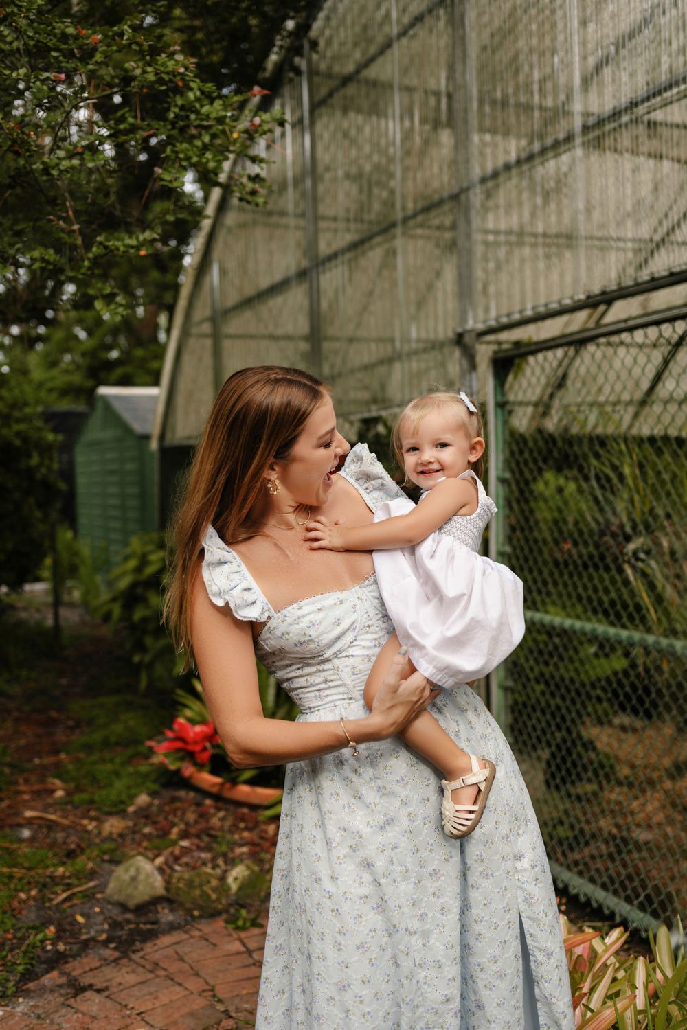 A woman in a light floral dress holds a smiling toddler in a white outfit beside a greenhouse surrounded by lush garden greenery.