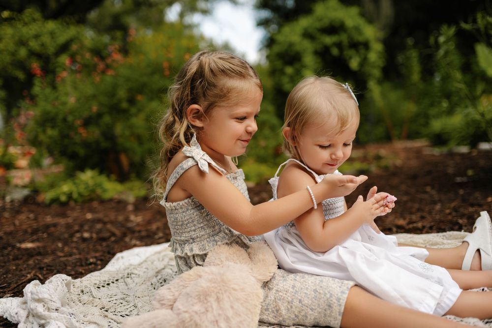 Two young girls in light summer dresses sit together on a blanket in a sunny garden, sharing a gentle moment with a plush teddy bear nearby.