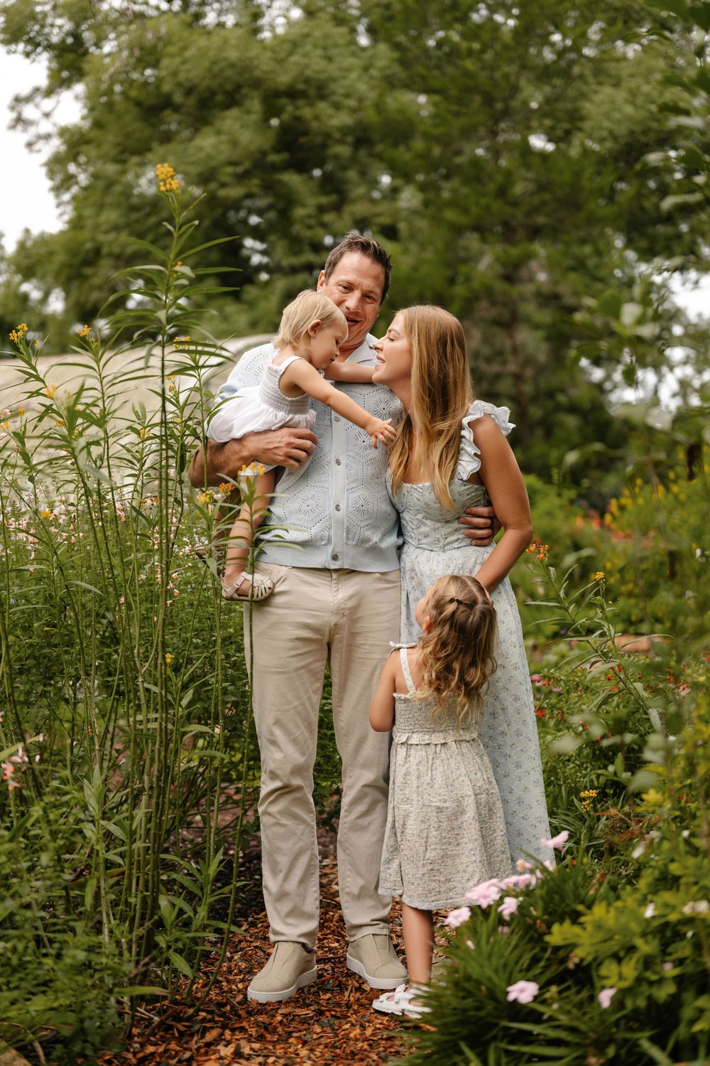 Smiling family of four embracing on a mulch path in a lush summer garden with pink flowers, a dad holding a toddler and a daughter nearby.