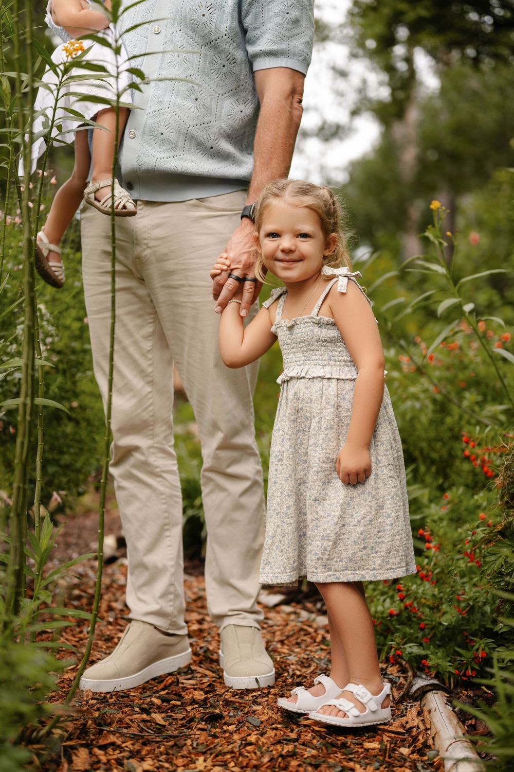 A smiling little girl in a gray smocked sundress holds an adult's hand while walking along a mulch garden path surrounded by greenery.
