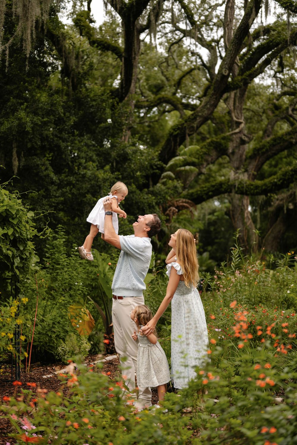 Family of four enjoying an outdoor portrait session in a lush garden with orange flowers, as a father lifts a baby beneath mossy oak trees.