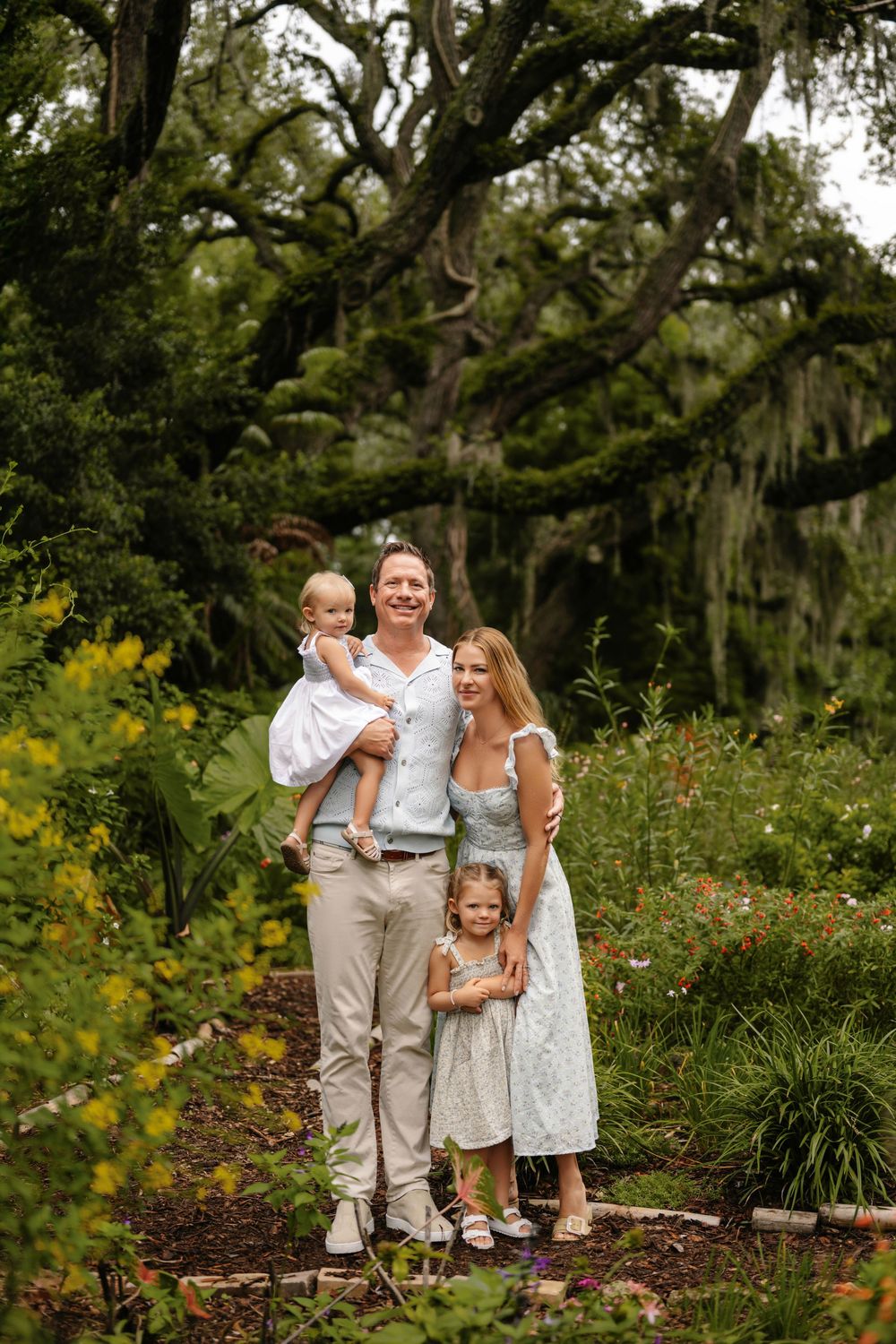 Family of four in light summer outfits posing together in a lush garden beneath large live oak trees draped with Spanish moss.