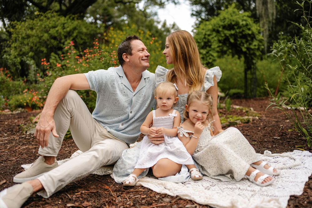 Smiling family of four sitting on a white blanket in a lush garden, with a father, mother and two young daughters in light summer outfits.