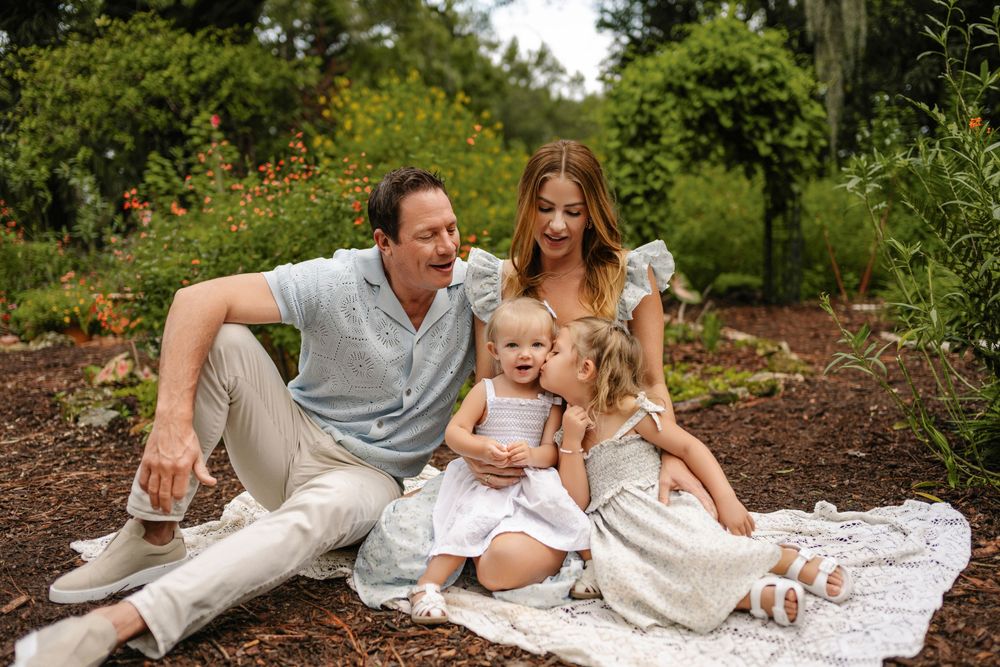 Smiling family of four sitting together on a blanket in a lush green garden, with two toddler girls in white dresses cuddling their parents.
