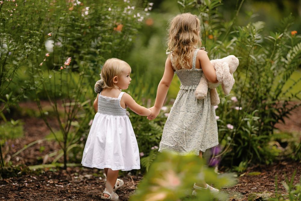 Two little girls in white and gray dresses hold hands while walking through a lush garden, one carrying a pink stuffed animal.
