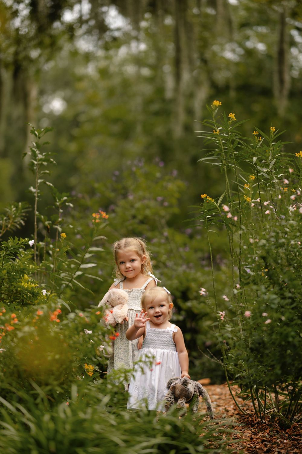 Two young girls in light dresses hold stuffed animals while standing on a mulch path surrounded by wildflowers in a sunlit garden.