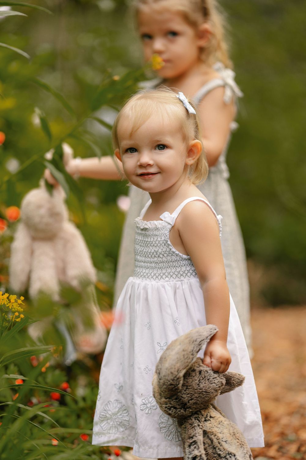 Two young girls in white smocked dresses stand in a sunny garden, one smiling and holding a plush bunny toy among colorful flowers.