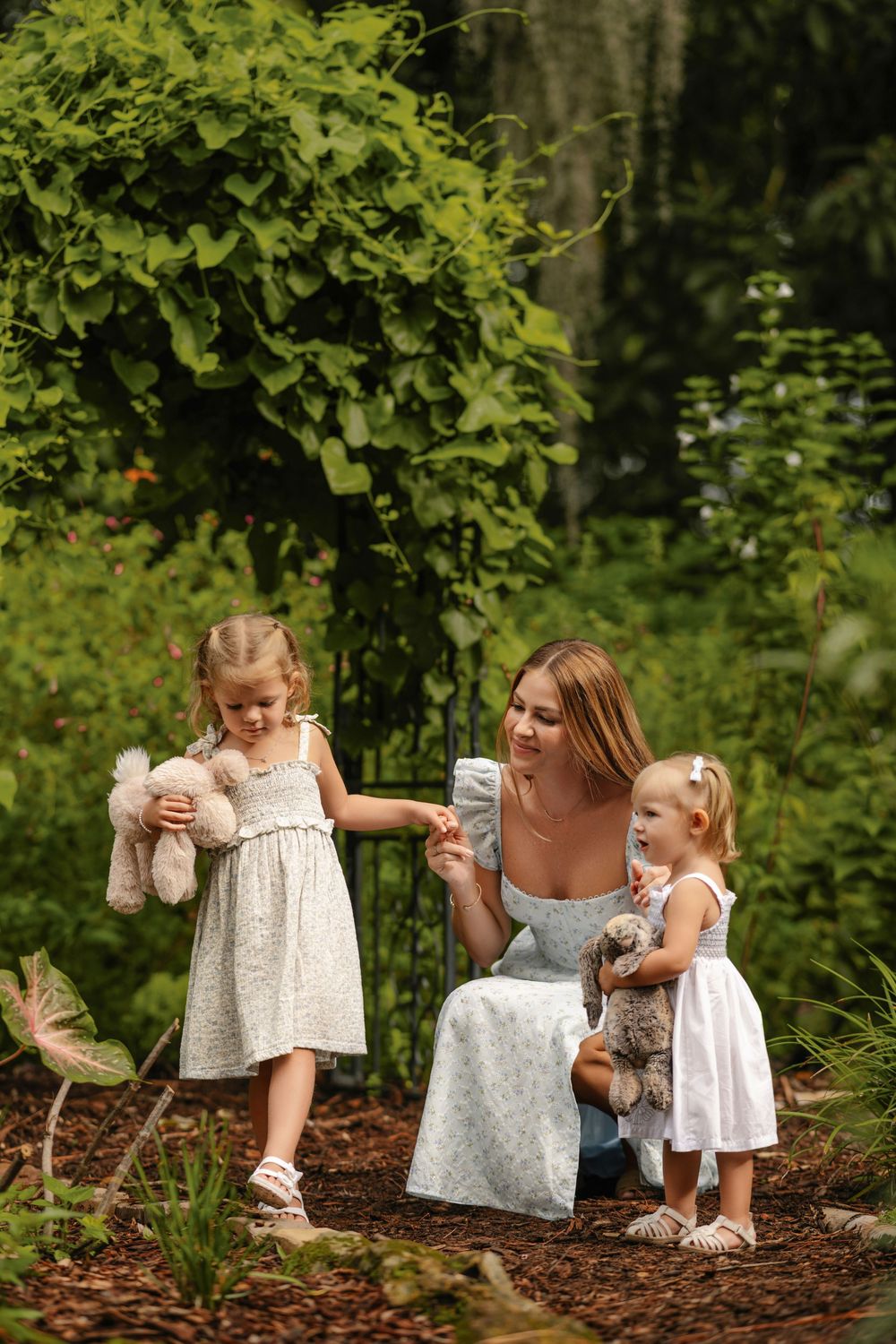 Mother in a blue dress sits in a lush garden holding hands with two little girls in white dresses, each carrying a stuffed animal.
