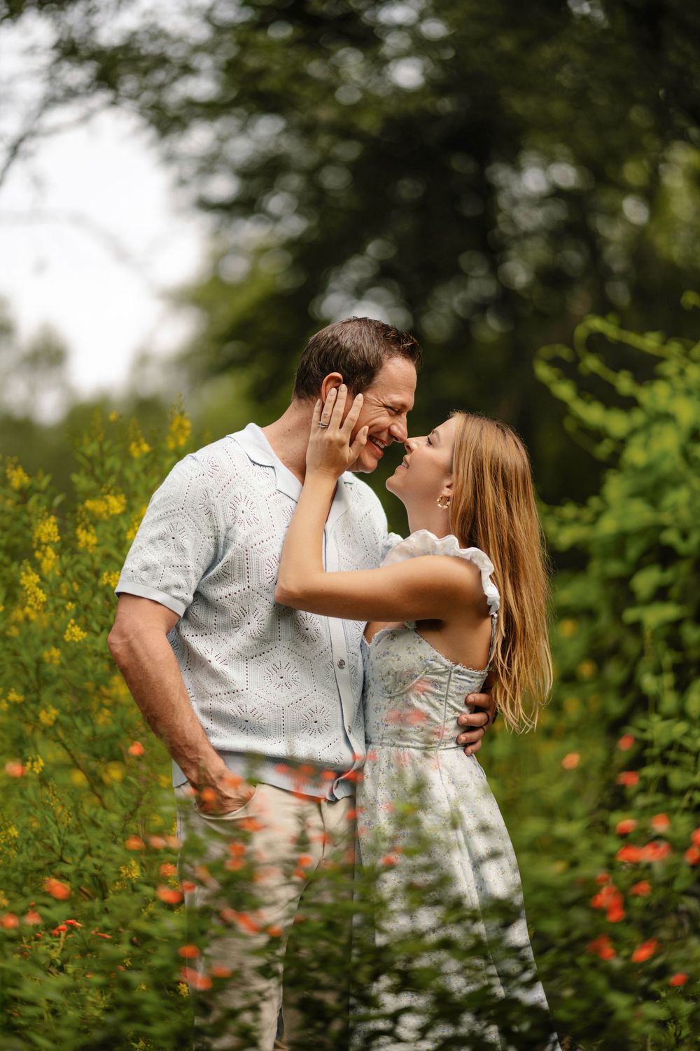 Smiling couple in an affectionate pose among green foliage and orange wildflowers during an outdoor engagement photo session.