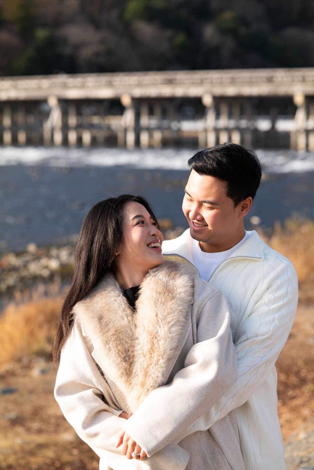 Smiling couple in matching cream coats embracing outdoors by a river with a wooden bridge and golden autumn grass behind them.