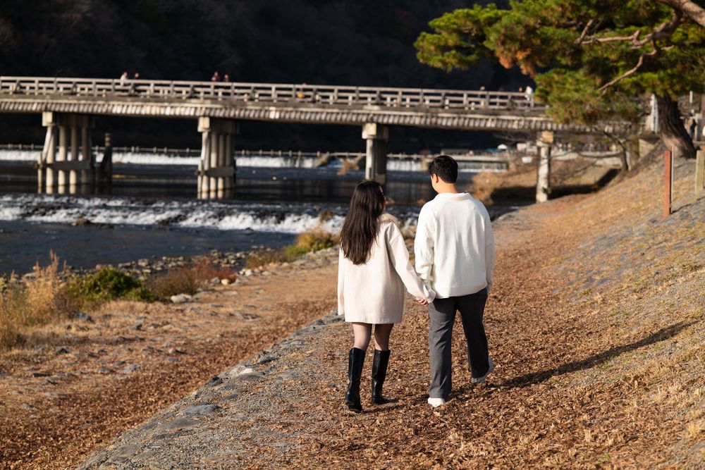 Couple in light jackets walk hand in hand along a leaf-covered riverside path toward a long wooden bridge on an autumn afternoon.