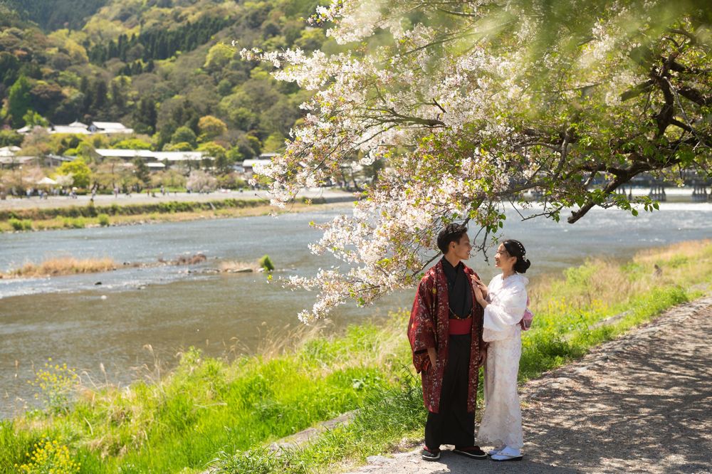 Couple in traditional Japanese kimono embracing beneath a blooming cherry blossom tree beside a calm river with forested mountains behind.