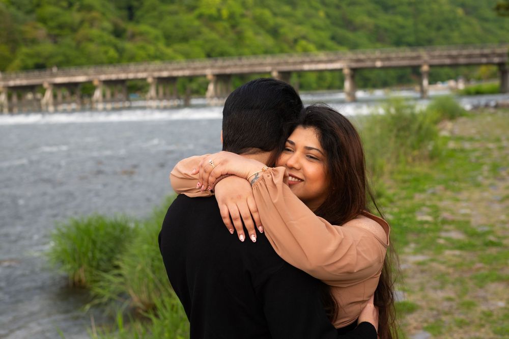 A smiling woman in a tan top embraces a man in a black shirt beside a calm river with a long bridge and green hills behind.