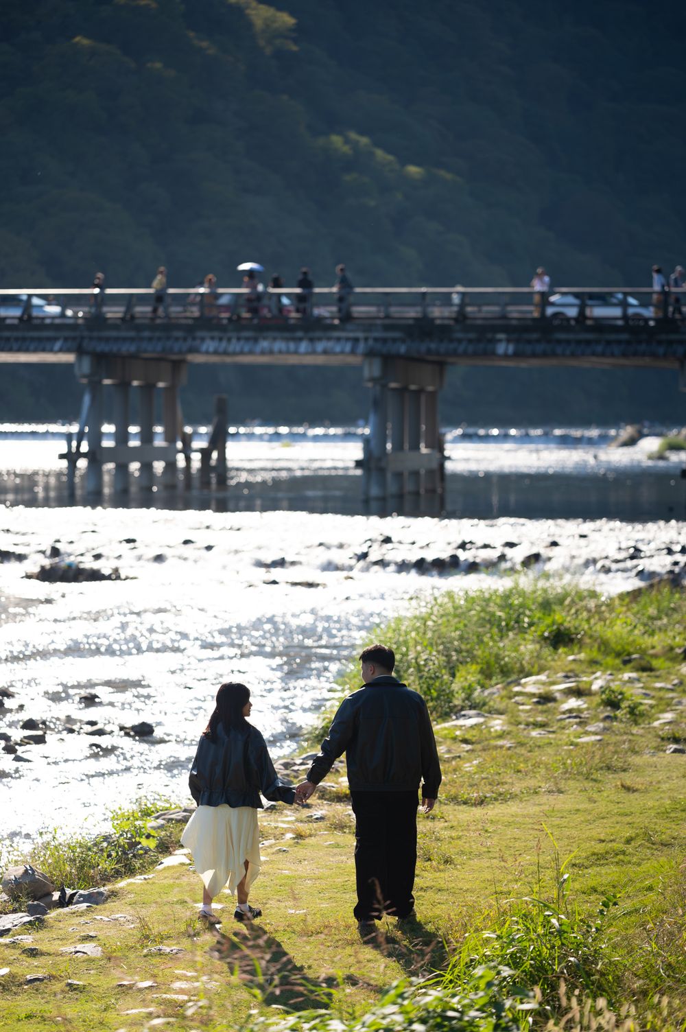 Couple holding hands walks along a grassy riverbank toward a wooden bridge with pedestrians, with forested hills and sunlit rapids beyond.