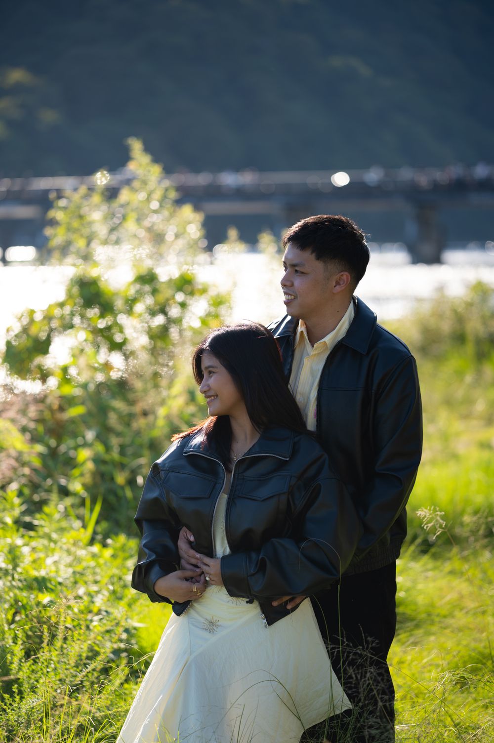 A couple in black leather jackets embraces on a grassy riverbank, with a bridge and sunlit water glowing softly in the background.
