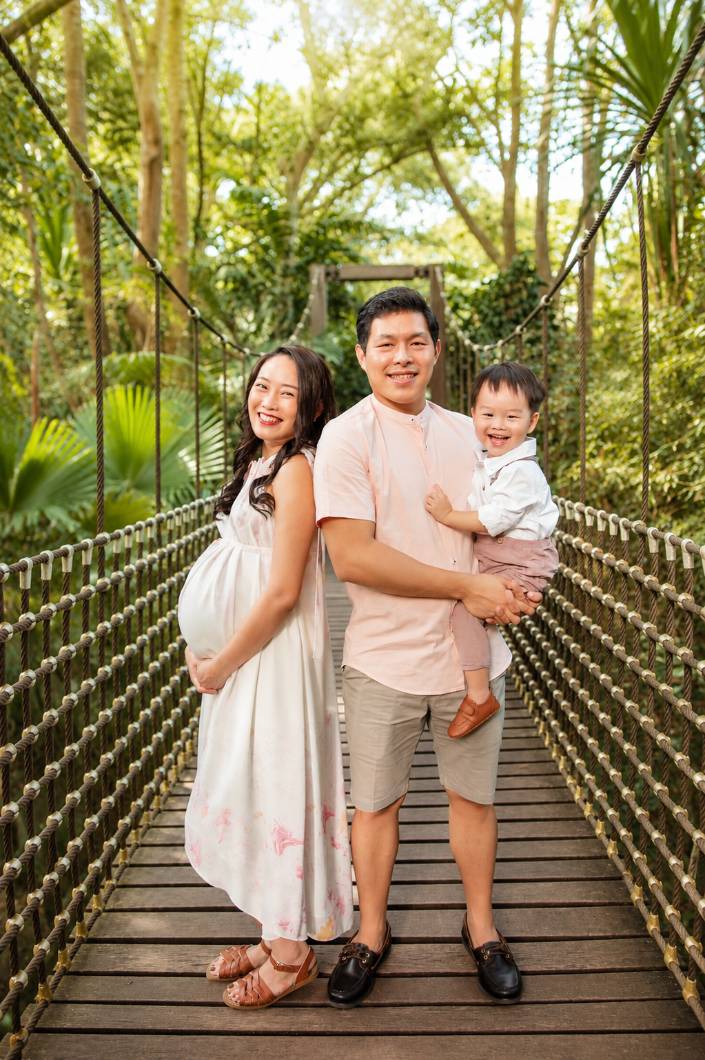 Smiling Asian family with pregnant mother and father holding toddler on a wooden suspension bridge.