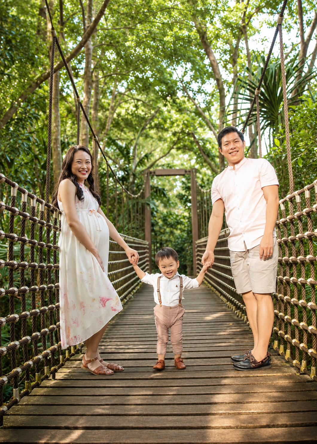 Asian family holding hands on a wooden suspension bridge surrounded by lush green tropical trees.