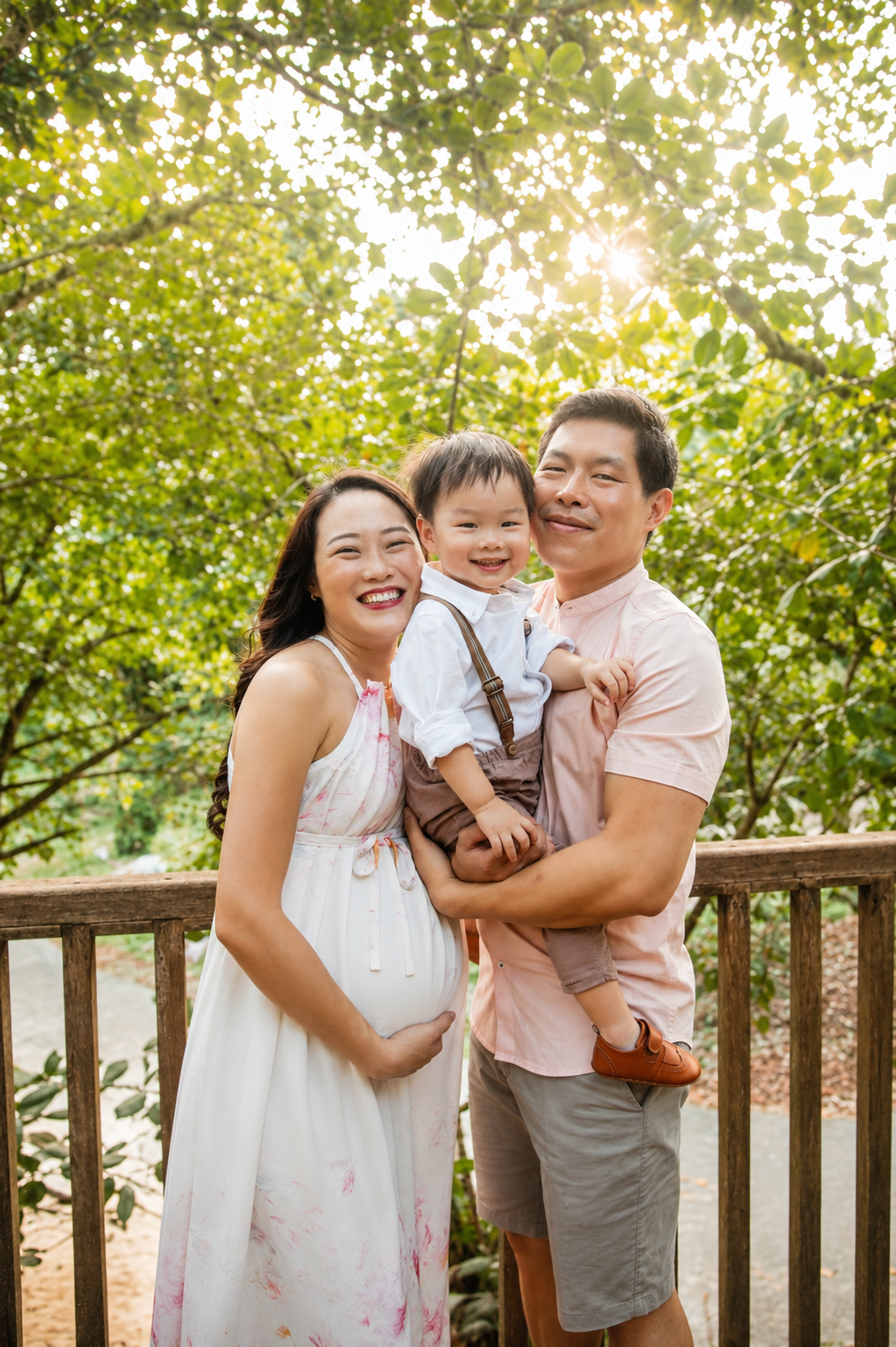 Asian family portrait with laughing pregnant mother, father holding toddler son on a wooden deck.