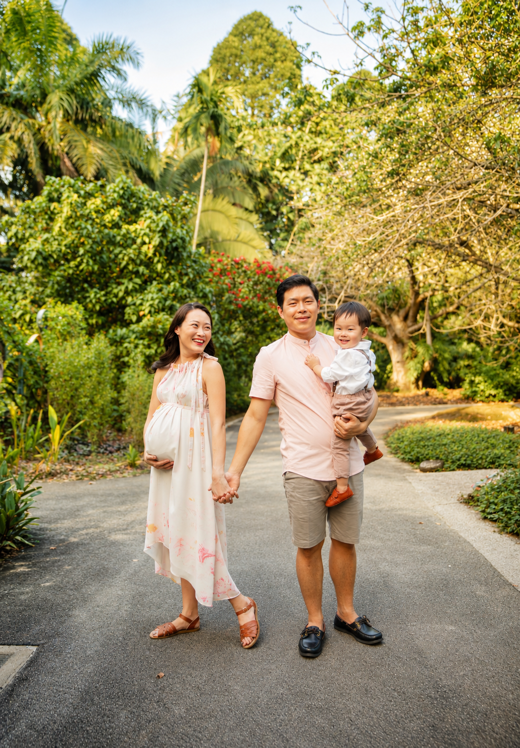 Joyful Asian family with pregnant mother, father carrying toddler, walking on a sunny garden path.